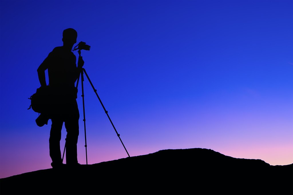 Silhouette of a photographer and tripod standing on top of a hill. It is sunset or sunrise and the sky is clear with a bold colour gradient from blue to violet to yellow.